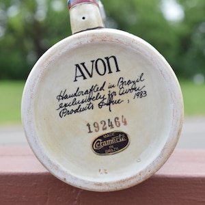 May include: The circular base of a ceramic stein features the text AVON, Handcrafted in Brazil exclusively for Avon Products Inc., 1983, and the number 192464. A black and gold sticker reads Made Ceramarte in Brazil at the bottom of the base.