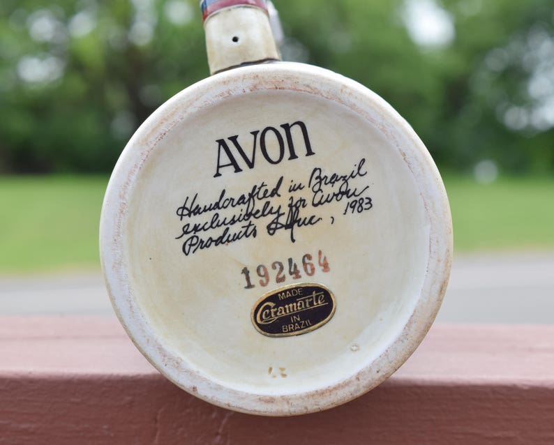 May include: The circular base of a ceramic stein features the text AVON, Handcrafted in Brazil exclusively for Avon Products Inc., 1983, and the number 192464. A black and gold sticker reads Made Ceramarte in Brazil at the bottom of the base.