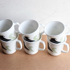 May include: Six white ceramic mugs with a black and green wave design. Each mug has a curved handle and a simple, clean aesthetic. The mugs are stacked in pairs, showcasing the design.