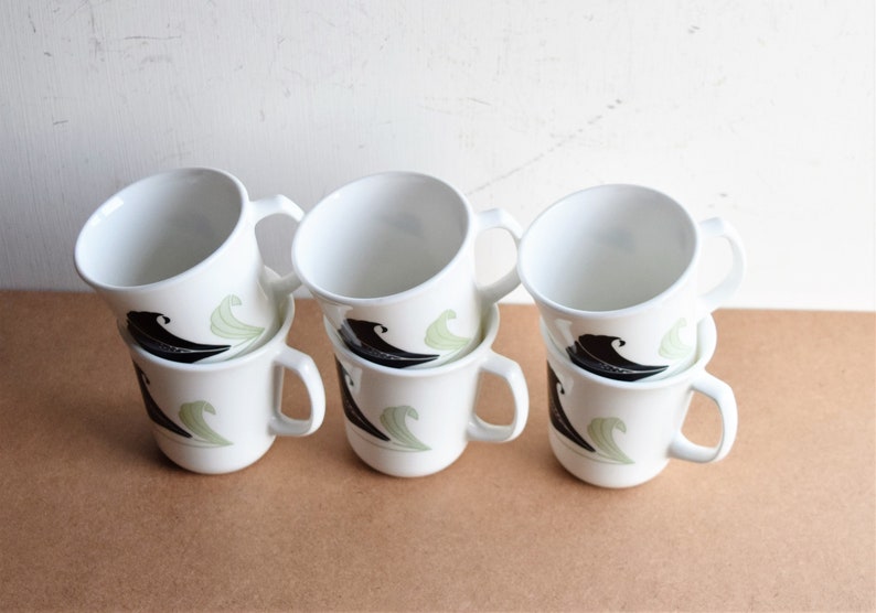 May include: Six white ceramic mugs with a black and green wave design. Each mug has a curved handle and a simple, clean aesthetic. The mugs are stacked in pairs, showcasing the design.