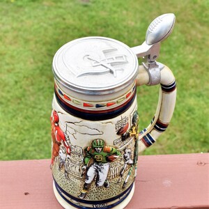 May include: A ceramic beer stein featuring a relief design of American football players in action on a field. The lid is metallic silver with a football-shaped thumb lift and a helmet emblem. The year 1960 is printed near the base of the stein.