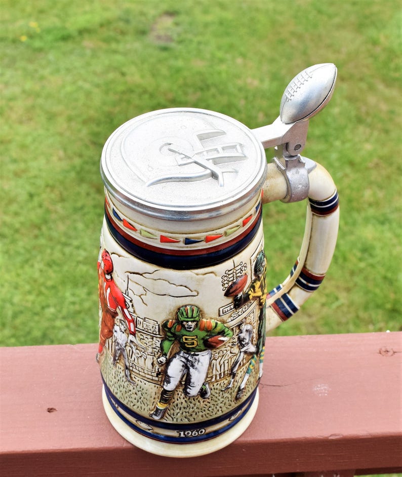 May include: A ceramic beer stein featuring a relief design of American football players in action on a field. The lid is metallic silver with a football-shaped thumb lift and a helmet emblem. The year 1960 is printed near the base of the stein.