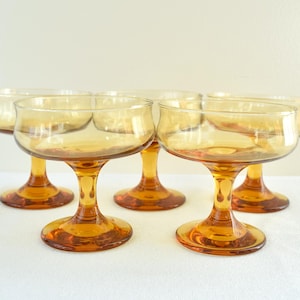 Op de afbeelding: Zes amberkleurige champagne coupe glazen. De glazen hebben een afgeronde komvorm, een steel en een cirkelvormige voet. Ze zijn gerangschikt op een wit oppervlak, met een neutrale achtergrond.