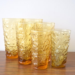 May include: A set of six amber-colored drinking glasses with a textured, bubbly surface. The glasses are of varying heights and shapes, arranged on a wooden surface against a white background. The glassware has a vintage aesthetic.