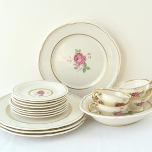 May include: A collection of vintage porcelain tableware. The set includes plates, saucers, and teacups, all with a cream-colored base, gold trim, and a pink rose motif. The plates have a scalloped edge.