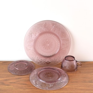 Pode incluir: Um conjunto de lou&ccedil;as de vidro rosa vintage, incluindo um prato grande, dois pratos menores e uma x&iacute;cara de ch&aacute;. Os pratos e a x&iacute;cara apresentam um design floral em relevo. As lou&ccedil;as est&atilde;o dispostas em uma superf&iacute;cie de madeira.
