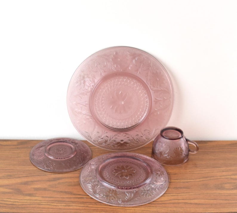 Pode incluir: Um conjunto de lou&ccedil;as de vidro rosa vintage, incluindo um prato grande, dois pratos menores e uma x&iacute;cara de ch&aacute;. Os pratos e a x&iacute;cara apresentam um design floral em relevo. As lou&ccedil;as est&atilde;o dispostas em uma superf&iacute;cie de madeira.