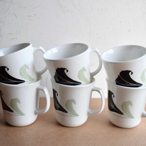 May include: Six white ceramic mugs stacked in two rows. Each mug features a black wave design and a light green leaf design. The mugs have a simple, curved handle and a slightly angled rim.