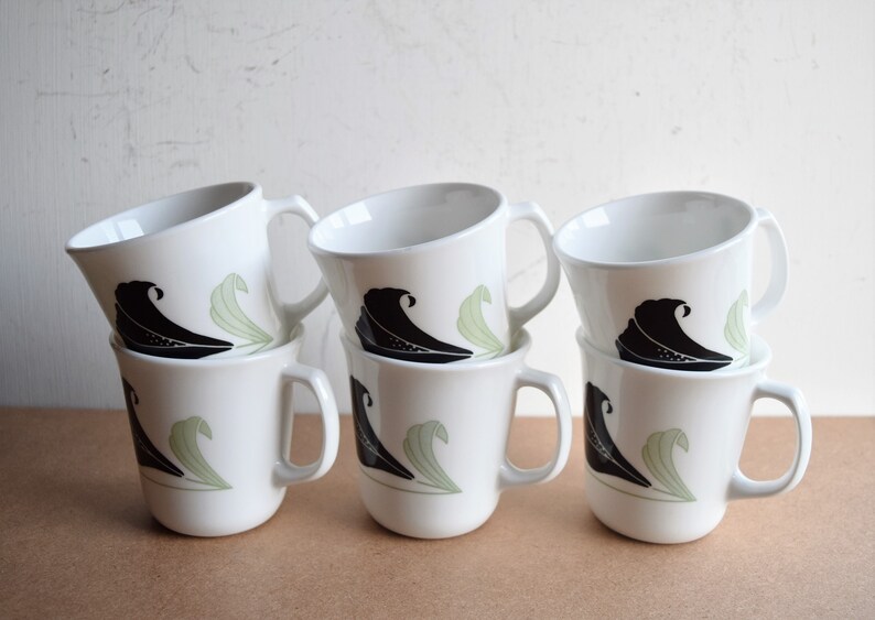 May include: Six white ceramic mugs stacked in two rows. Each mug features a black wave design and a light green leaf design. The mugs have a simple, curved handle and a slightly angled rim.
