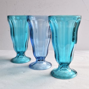 May include: Three blue glass sundae glasses with a fluted design. The glasses are arranged in a row on a white surface.