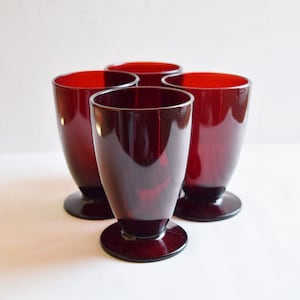 May include: Four ruby red stemmed glasses. The glasses have a flared shape and a solid base. The interior of the glasses is a bright red, while the exterior appears a darker shade of red. The glasses are arranged in a group.