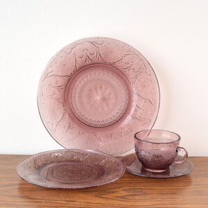 Pode incluir: Um conjunto de vidro vintage lil&aacute;s, incluindo um prato grande, um prato pequeno, uma ch&aacute;vena de ch&aacute; e um pires. As pe&ccedil;as apresentam um design floral em relevo. A loi&ccedil;a de vidro &eacute; exibida numa superf&iacute;cie de madeira.
