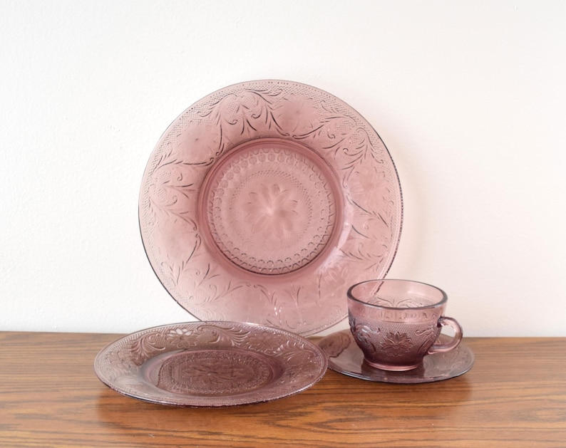 Pode incluir: Um conjunto de vidro vintage lil&aacute;s, incluindo um prato grande, um prato pequeno, uma ch&aacute;vena de ch&aacute; e um pires. As pe&ccedil;as apresentam um design floral em relevo. A loi&ccedil;a de vidro &eacute; exibida numa superf&iacute;cie de madeira.