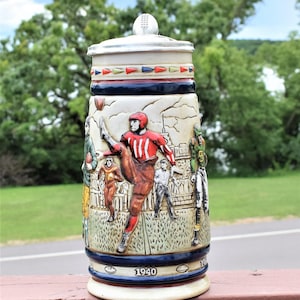May include: A ceramic beer stein featuring a relief design of American football players in action. The stein has a metal lid with a football finial, a decorative border of colored triangles, and the year 1940 printed on the base in dark blue.