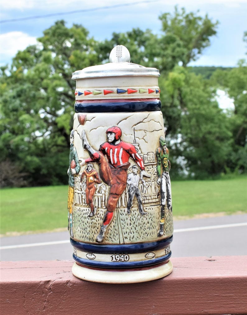 May include: A ceramic beer stein featuring a relief design of American football players in action. The stein has a metal lid with a football finial, a decorative border of colored triangles, and the year 1940 printed on the base in dark blue.