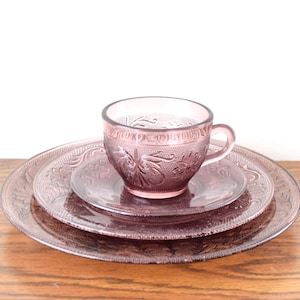 Pode incluir: Um conjunto de ch&aacute; vintage em vidro tingido de rosa. A ch&aacute;vena, o pires pequeno e o prato apresentam um design floral em relevo. O conjunto &eacute; exibido numa superf&iacute;cie de madeira sobre um fundo branco.