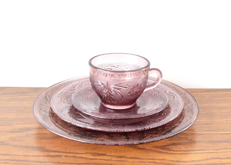 Pode incluir: Um conjunto de ch&aacute; vintage em vidro tingido de rosa. A ch&aacute;vena, o pires pequeno e o prato apresentam um design floral em relevo. O conjunto &eacute; exibido numa superf&iacute;cie de madeira sobre um fundo branco.