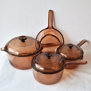 Puede incluir: Un juego de utensilios de cocina de vidrio ámbar vintage, que incluye ollas con tapas y una sartén. Los utensilios de cocina están colocados sobre una superficie blanca, mostrando su diseño transparente y estilo clásico. El juego incluye varios tamaños.