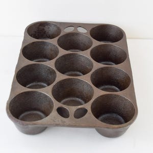 May include: A vintage cast iron muffin tin with twelve round cups. The pan is dark brown with a slightly textured surface and is designed for baking muffins or cupcakes. The tin has a rectangular shape with rounded corners and small feet.
