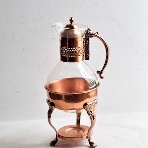 May include: A vintage copper and glass coffee maker. The clear glass carafe sits within a copper frame and is supported by ornate legs. The handle and lid are also copper, creating a cohesive, antique aesthetic.