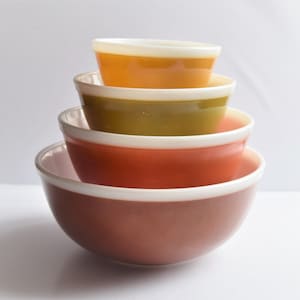 May include: A stack of four vintage mixing bowls in shades of orange, green, red-orange, and brown. Each bowl has a white rim. The bowls are nested inside each other, with the largest bowl at the bottom.