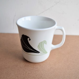May include: White ceramic mug with a curved handle. The mug features a black and light green wave design on the side. The mug is empty and sits on a brown surface.