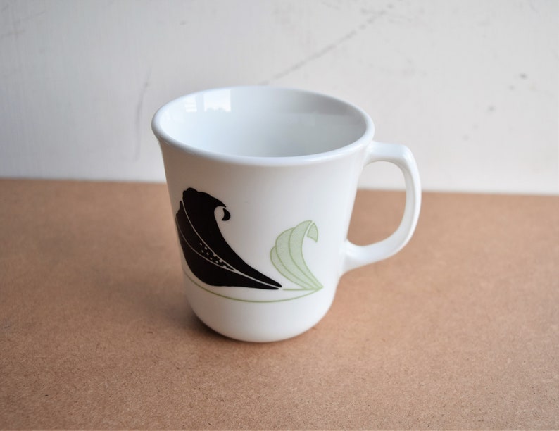 May include: White ceramic mug with a curved handle. The mug features a black and light green wave design on the side. The mug is empty and sits on a brown surface.