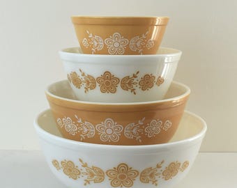 Puede incluir: Un conjunto de cuatro cuencos de mezcla vintage apilados. Los cuencos alternan entre blanco y ámbar, cada uno adornado con un diseño floral y de hojas doradas. Los cuencos están anidados, mostrando sus tamaños graduados. Medidas en centímetros.