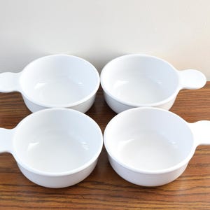 May include: Four white ceramic bowls with handles are arranged on a wooden surface. The bowls are round with a small, rounded handle on one side. The bowls are empty and have a glossy finish.