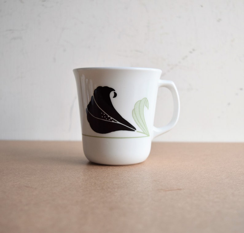 May include: White ceramic mug with a black floral design and a green accent line. The mug features a curved handle and a rounded base. The floral design is stylized and appears on the front of the mug.