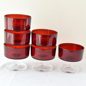 Op de afbeelding: Zes robijnrode glazen dessertkommen met heldere stelen zijn gerangschikt op een wit oppervlak. De kommen hebben een cilindrische vorm en zijn gestapeld in groepen van drie en twee, met één kom alleen.