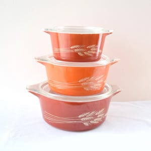 Könnte beinhalten: Ein Stapel von drei Vintage Pyrex Auflaufformen in Orange- und Rottönen. Jede Form hat einen Deckel aus klarem Glas und ein weißes Weizenähren-Design. Die Formen sind übereinander gestapelt.