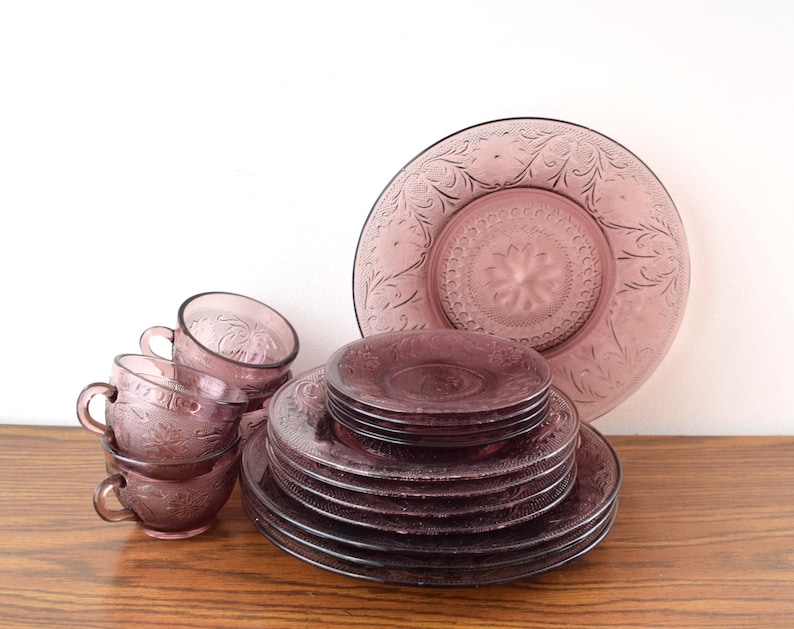 Pode incluir: Uma cole&ccedil;&atilde;o de lou&ccedil;as de vidro roxo vintage, incluindo x&iacute;caras de ch&aacute;, pires e pratos. Os pratos e x&iacute;caras apresentam um design floral em relevo. O conjunto inclui um prato grande, v&aacute;rios pratos menores e uma pilha de x&iacute;caras de ch&aacute;.