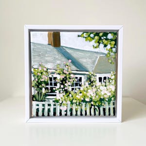 May include: An original painting of a house with a white picket fence, windows, and a chimney. The house is painted in shades of gray and white, with green and pink flowers and foliage. The artwork is framed in a white square frame.