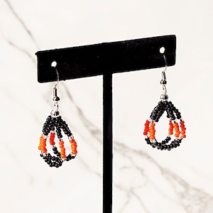 May include: A pair of dangle earrings with black and orange beads. The earrings are hanging from a black display stand.