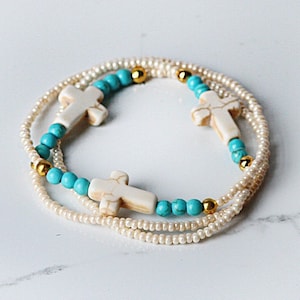 May include: A white and turquoise beaded bracelet with three white cross charms. The bracelet is made of small white beads and turquoise beads.