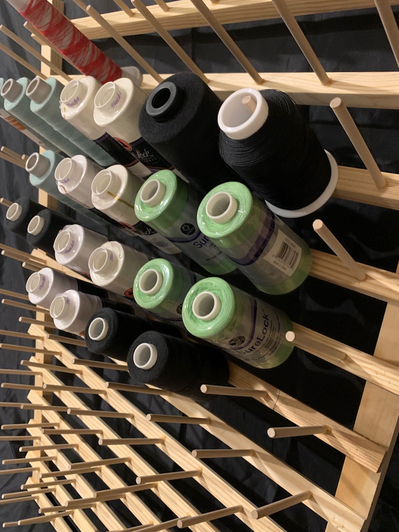 May include: A wooden yarn rack with spools of thread in various colors, including black, white, green, and red. The rack has multiple rows of dowels for holding the spools.