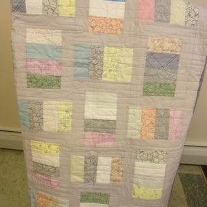 Könnte beinhalten: Eine handgefertigte Steppdecke mit hellgrauem Hintergrund und rechteckigen Flicken in verschiedenen Pastellfarben. Die Flicken umfassen weiße, rosa, gelbe, blaue und grüne Stoffe mit gemusterten und unifarbenen Designs. Die Steppdecke hat ein gestepptes Muster.