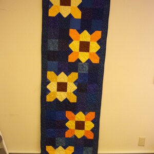 May include: A blue quilted table runner with five yellow, orange, and brown sunflower designs.
