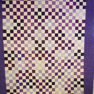 May include: A purple and white checkered quilt with a purple border. The quilt is made of squares of different shades of purple and white.
