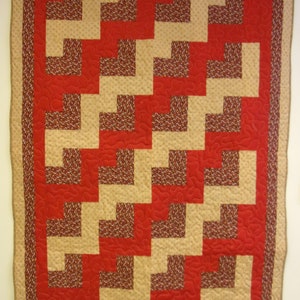 May include: A handmade quilt with a geometric pattern. The quilt features a red background with beige and patterned brown blocks arranged in a repeating design. The border is beige with a patterned brown trim.