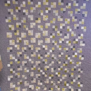 May include: A quilt made of small squares in shades of gray, white, and yellow. The squares are arranged in a checkerboard pattern. The quilt is quilted with a wavy pattern.
