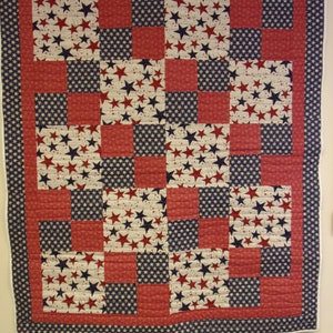 Puede incluir: Una colcha de retazos roja, blanca y azul con un patrón de estrellas. La colcha está hecha de cuadrados de diferentes telas y está cosida en un patrón de cuadrícula.