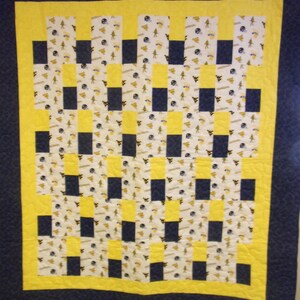 May include: A handmade quilt with a navy blue border and a yellow and white checkered pattern. The quilt's center features alternating blocks of yellow and navy blue squares, with a white background filled with small, patterned images. The quilt is likely for a bed or wall decor.
