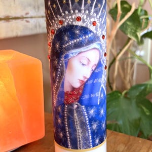 May include: A tall cylindrical glass prayer candle featuring a printed image of a figure wearing a blue head covering and red garment. The image is decorated with small red and gold circular embellishments arranged in a halo pattern around the head.