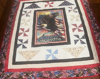 May include: A red, white, and blue quilt with a patriotic theme. The quilt features a bald eagle with outstretched wings, a red, white, and blue American flag, and the words "America The Beautiful". The quilt is made with a variety of fabrics, including stars, pinwheels, and a brown fabric with a white polka dot pattern.