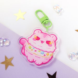 Puede incluir: Un llavero de un personaje de dibujos animados rosa y blanco con un cierre de metal verde. El personaje tiene un cuerpo rosa, pelaje blanco y una bufanda a rayas rosa y blanca. El personaje tiene una expresión feliz con los ojos cerrados y un corazón rosa en el pecho.