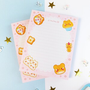 May include: Two pink and white lined notepads with a cute bear and food theme. The notepads have a white lace border and feature illustrations of bears wearing hats and holding food like pizza, cupcakes, and toast. The text "Sweet Bakery" is printed on the top of the notepad.
