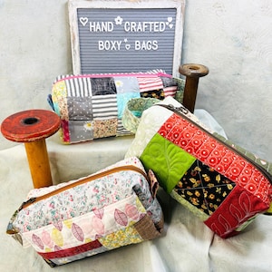 May include: Three rectangular quilted boxy bags with zipper closures. One bag features a patchwork of gray, white, and floral squares. Another has red, green, and black floral patchwork. The third has a floral print with leaf patterns. A sign reads HAND CRAFTED BOXY BAGS.