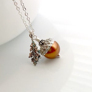 Puede incluir: Collar plateado con un colgante de bellota. La bellota tiene un cuerpo marrón perlado y una tapa plateada, acompañada de un dije de hoja plateada. La cadena es delicada y el colgante está centrado.